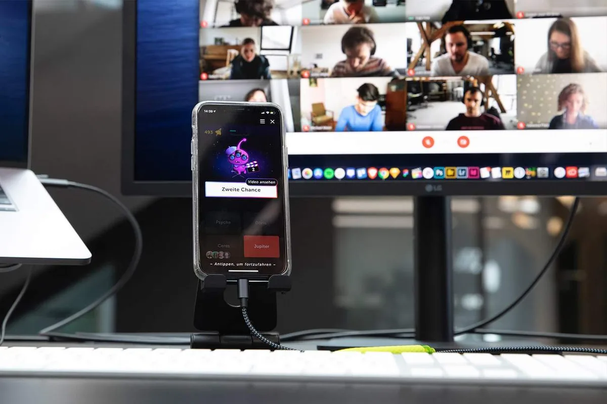 A phone on a stand in front of a monitor: a Lotum quiz game on the phone, colleagues in a video call on the screen behind it.